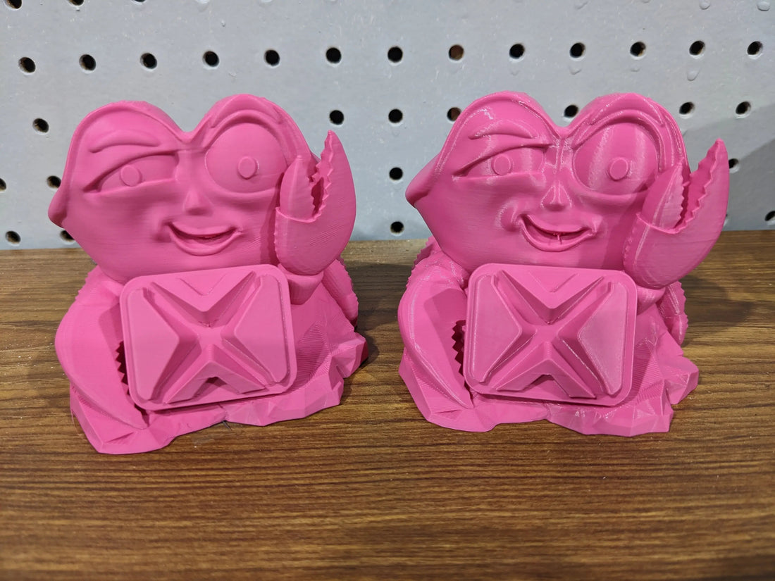 Two pink 3D printed crab figurines on a wooden surface with a pegboard background.